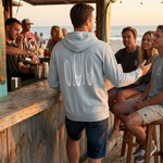 man at a beach bar wearing a surfboard sweatshirt with his friends on the beach